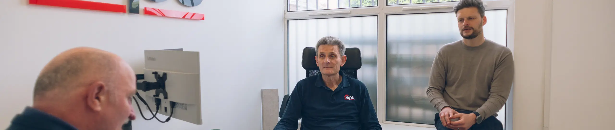 Two men in an office by a window, one seated in an eps polo shirt and one sitting on the windowsill, with a third man's head out of focus in the foreground